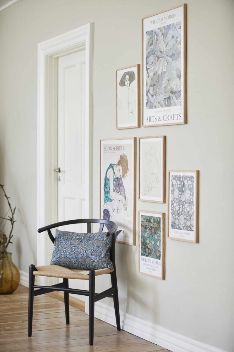William Morris pude Daisy 50x33 siddende på en stol foran en væg med billeder på